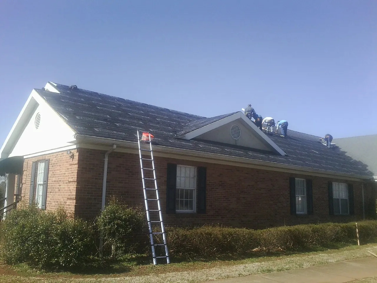Crew tearing shingles off a brick ranch roof with a ladder set — Metal Roof Repair work by our crew in Rexburg