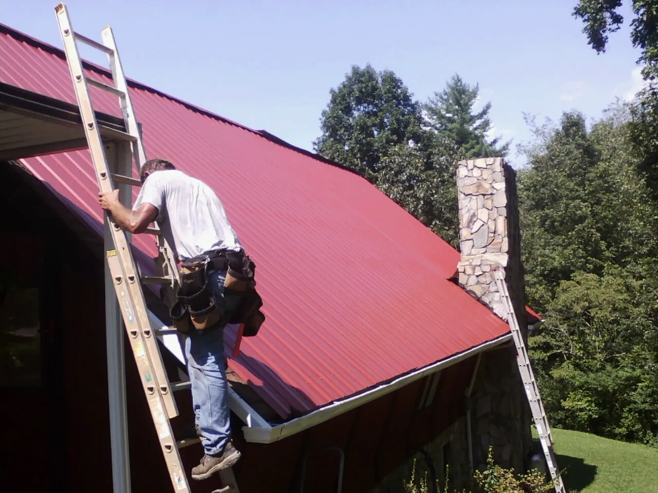 Roofer on a ladder beside a red metal roof and stone chimney — Soffit & Fascia Repair by Roofing Brothers in Rexburg
